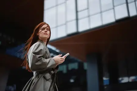Стильная деловая женщина с телефоном в руке, одетая в модную одежду, прогуливается по городу рядом с офисными башнями