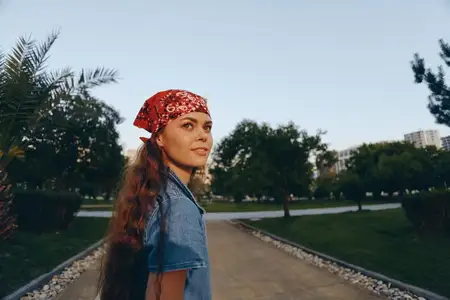 Женщина в стиле casual, одетая в современный деним в стиле бохо-вестерн с красной банданой, стоит на улице в солнечный день в окружении зелени и городских элементов