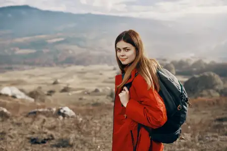 Рыжеволосая женщина в куртке с рюкзаком путешествует, занимается пешим туризмом в горах, дышит свежим воздухом
