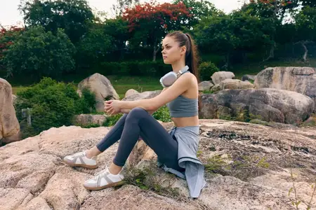 Подтянутая молодая женщина в спортивной одежде сидит на скалистой местности, погруженная в природу с наушниками, передавая темы фитнеса, расслабления и заботы о здоровье