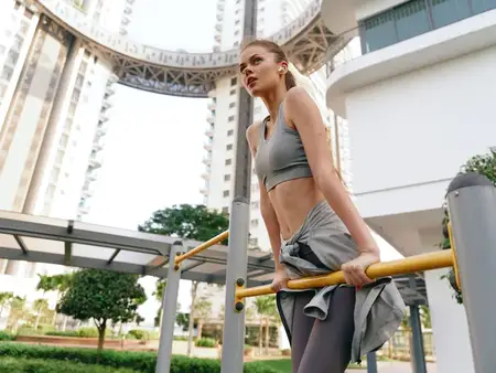 Спортивная женщина занимается на открытом воздухе, сосредоточившись на подтягиваниях, в окружении современной архитектуры и зелени в яркой городской среде