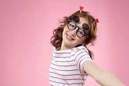 Маленькая девочка в модных очках и с зажимами для волос, её эмоции на розовом фоне с сердечками