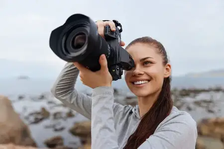 Счастливая молодая женщина в серой толстовке профессионально фотографирует красивый морской пейзаж, воплощая любовь к фотографии и природе