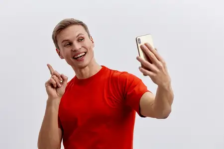 Улыбающийся мужчина в красной футболке делает селфи на смартфон для видеочата или общения в социальных сетях, выражая счастье и вовлеченность в технологии цифровой коммуникации на белом фоне