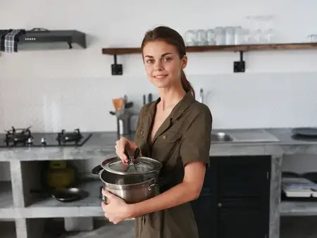 Женщина на стильной кухне готовит еду, держит в руках кастрюлю для приготовления ужина и улыбается, показывая зубы