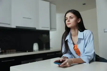 Обеспокоенная женщина сидит за кухонным столом с мобильным телефоном в руке, выражая тревогу и беспокойство
