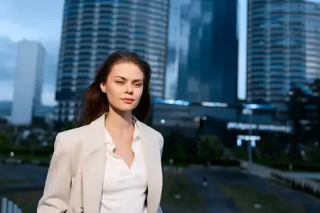 Уверенная бизнес-леди в белом блейзере и рубашке позирует на улице на фоне современных городских небоскребов в сумерках