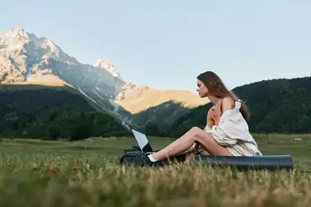 Женщина работает удаленно на солнечном лугу, ноутбук открыт на траве, в окружении гор и голубого неба, демонстрируя баланс между продуктивностью и спокойствием на природе