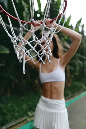 Юная спортсменка в белом кроп-топе и юбке демонстрирует силу и сосредоточенность, тренируясь в баскетболе на фоне пышной зелени, передавая энергию и летнее настроение