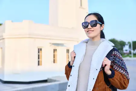 Уличная сцена представляет стильную женщину в пальто и куртке, в солнцезащитных очках, демонстрирующую моду, стиль и архитектуру с уверенной позой на фоне солнечного городского пейзажа