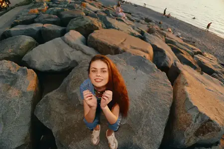 Женщина в стиле life style, одетая в современный бохо-вестерн деним стритстайл, радостно позирует на открытом воздухе на больших камнях у пляжа на закате, запечатленная в теплых кинематографических…