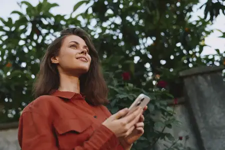 Молодая женщина на улице смотрит вверх, мягко улыбаясь, держа смартфон, в обрамлении зеленых листьев и цветов, красная рубашка и расслабленная поза передают естественную повседневную сцену