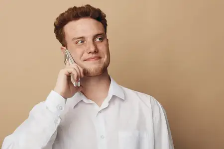 Улыбающийся молодой человек в белой рубашке держит смартфон у уха, глядя в сторону