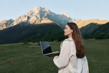 Человек использует ноутбук в природном ландшафте, сочетая работу и исследование среди открытого пространства, яркого света, далеких пиков и пересеченных холмов