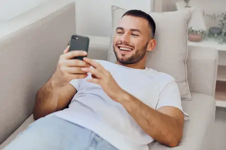 Улыбающийся мужчина, удобно устроившись на диване, занимается цифровым детоксом, просматривая смартфон