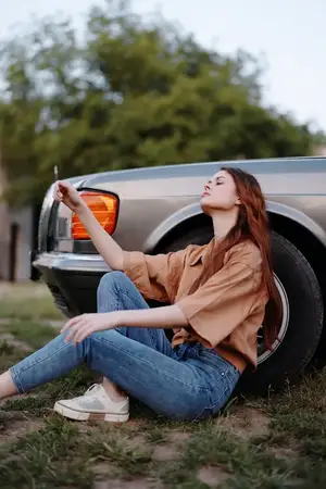 Расслабленная молодая женщина сидит рядом с винтажным автомобилем в джинсовых брюках и свободной рубашке, излучая спокойствие и радость в естественной природной обстановке