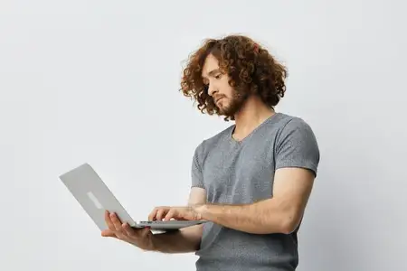 Молодой человек с кудрявыми волосами сосредоточенно работает за ноутбуком, одетый в серую футболку, в светлой минималистичной обстановке