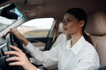 Женщина за рулем автомобиля: она уверенно сидит на водительском сиденье в белой рубашке, держа руки на рулевом колесе, и привлекательно смотрит в камеру, подчеркивая свои навыки вождения и…