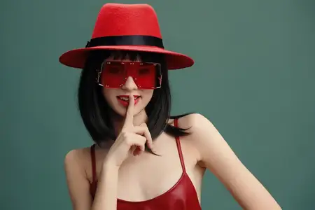 Азиатская женщина в красном платье и шляпе с солнцезащитными очками прикладывает палец к губам, стоя на зеленом фоне
