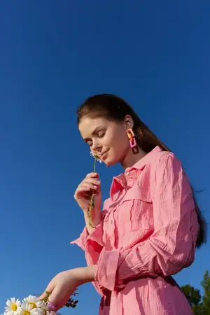Веселая молодая женщина в розовом платье держит ромашки и улыбается под ярким голубым небом