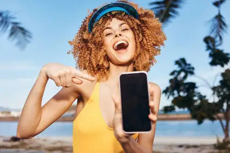 Счастливая женщина с кудрявыми волосами держит смартфон и указывает на экран; на заднем плане — пальмы и голубое небо