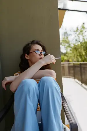 Она сидит, подтянув колени и обхватив их руками, глядя вдаль, в то время как мягкий естественный свет подчеркивает спокойный, простой момент на улице в солнечный день