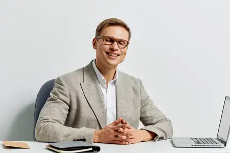 Улыбающийся мужчина европеоидной внешности в очках и сером пиджаке сидит за столом с ноутбуком, демонстрируя образ жизни цифрового кочевника и концепцию удаленной работы для бизнеса и…