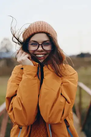 Уличная сцена отдыха: улыбающаяся женщина на улице зимой, очки и шапка-бини, оранжевое пальто, поправляет воротник, ветер в волосах, золотистый солнечный свет, малая глубина резкости, парк и город в…
