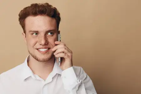 Счастливый рыжеволосый мужчина в белой рубашке, прижимающий смартфон к уху, смотрит в сторону с улыбкой
