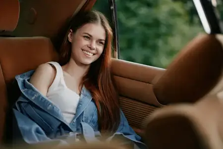 Улыбающаяся молодая женщина излучает радость, удобно устроившись в автомобиле, одетая в повседневный наряд с джинсовой курткой, воплощая весёлое и беззаботное настроение