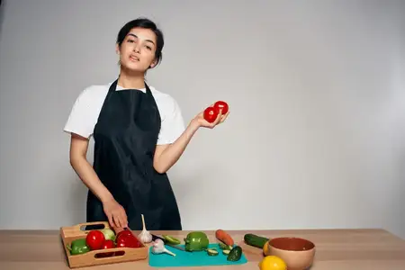 Домохозяйка в черном фартуке на кухне с овощами, свежей едой на светлом фоне