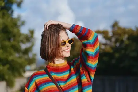 Женский портрет в радужном свитере и ретро-солнцезащитных очках, улыбающейся на улице в солнечном парке
