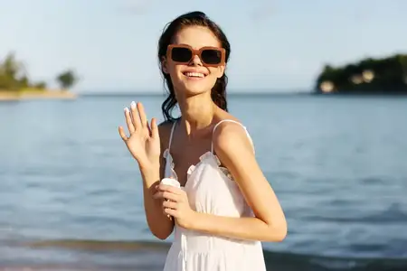 Улыбающаяся женщина в солнцезащитных очках машет рукой, держа солнцезащитный крем, на фоне моря и голубого неба, наслаждаясь летним пляжным днем, беззаботная и радостная атмосфера