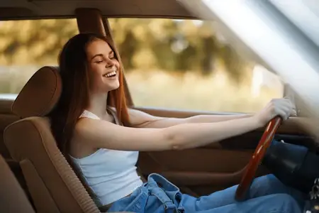 Веселая молодая женщина в повседневной одежде смеется за рулем, демонстрируя свое счастье и свободу в естественной обстановке, что идеально подходит для концепций, связанных с путешествиями…