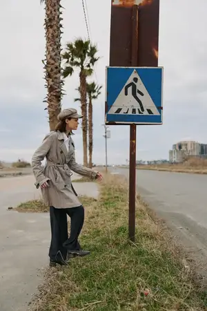 Молодая женщина в тренчкоте и шляпе рассматривает знак пешеходного перехода у пустой дороги, обсаженной пальмами