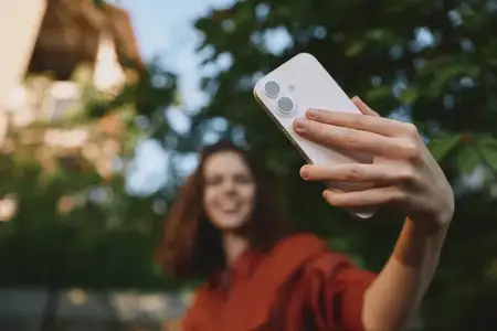 Молодая женщина с каштановыми волосами держит смартфон на вытянутой руке, чтобы сфотографировать себя на улице