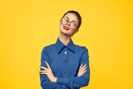 Женщина в очках и голубой рубашке жестикулирует руками на желтом фоне, обрезанный вид