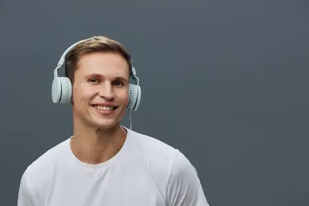 Улыбающийся молодой мужчина европеоидной расы в белых наушниках и белой футболке смотрит в камеру с позитивными эмоциями, изолированный на сером фоне