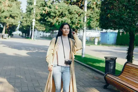 Уличная сцена: модная женщина в пальто, на шее висит камера, спокойная парковая дорожка, рядом скамейка