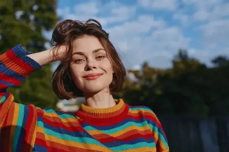 Женский портрет: улыбка, разноцветный свитер на улице, солнечный свет подсвечивает лицо, повседневная короткая стрижка, уверенный взгляд и непринужденная композиция, идеально подходящая для портрета…