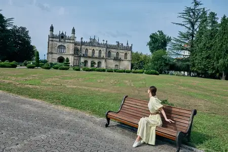 Уличная сцена показывает одинокую женщину на скамейке рядом с большим замком, передавая расслабление, природу и вневременное архитектурное очарование в спокойной обстановке