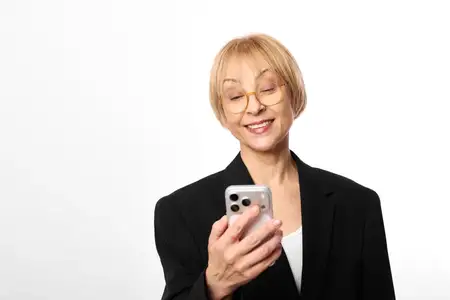 Улыбающаяся пожилая женщина в очках держит смартфон, портрет на белом студийном фоне