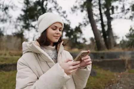 Молодая женщина в толстой куртке и вязаной шапке на улице в парке, листает телефон