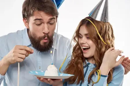 Энергичные мужчина и женщина с тортом и в праздничных колпаках отмечают день рождения на светлом фоне