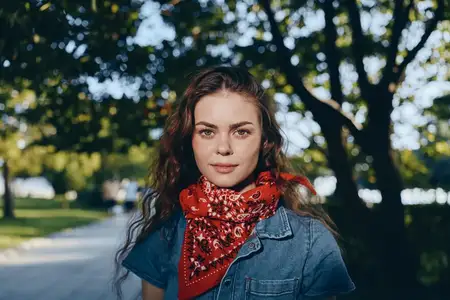 Женщина в стиле лайфстайл, одетая в современную бохо-вестерн джинсовую уличную моду с красной банданой, уверенно стоит в залитом солнцем парке, демонстрируя естественные пленочные цветовые тона и…