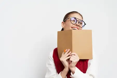Весёлая молодая женщина в очках прячет улыбку за коричневой книгой, излучая радость и энтузиазм
