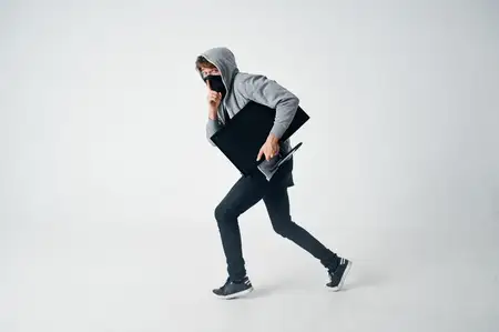 Мужчина в капюшоне, скрывающий лицо под маской, сидит перед монитором компьютера
