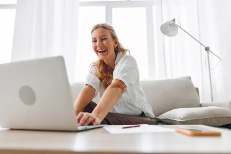 Счастливая женщина работает из дома на уютном диване с ноутбуком, наслаждаясь продуктивным и расслабленным днем