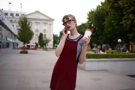 Женщина с короткой стрижкой на улице, в очках, со стаканом напитка в руке, идёт пешком