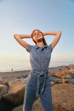 Женщина в стиле lifestyle, одетая в современный бохо-вестерн деним стритстайл, уверенно стоит на каменистом пляже на закате, улыбается, заложив руки за голову; плёночные цветовые тона усиливают…